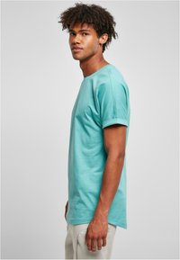 Camiseta de algodón turquesa con mangas enrolladas, cuello redondo y un ajuste relajado, combinada con pantalones de color claro. Diseño liso sin patrones.