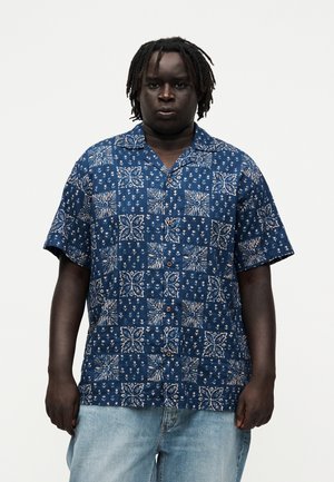 Homme à la peau foncée et aux dreadlocks portant une chemise bleue à manches courtes boutonnée avec des motifs floraux blancs et un jean bleu clair.