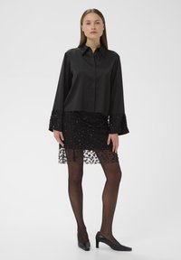 Crna bluzica s oštrim ovratnikom i dugim rukavima, u kombinaciji s crnom suknjom ukrašenom perlicama koja ima čipkastu nadogradnju i crnim tajicama, upotpunjena visokim potpeticama.