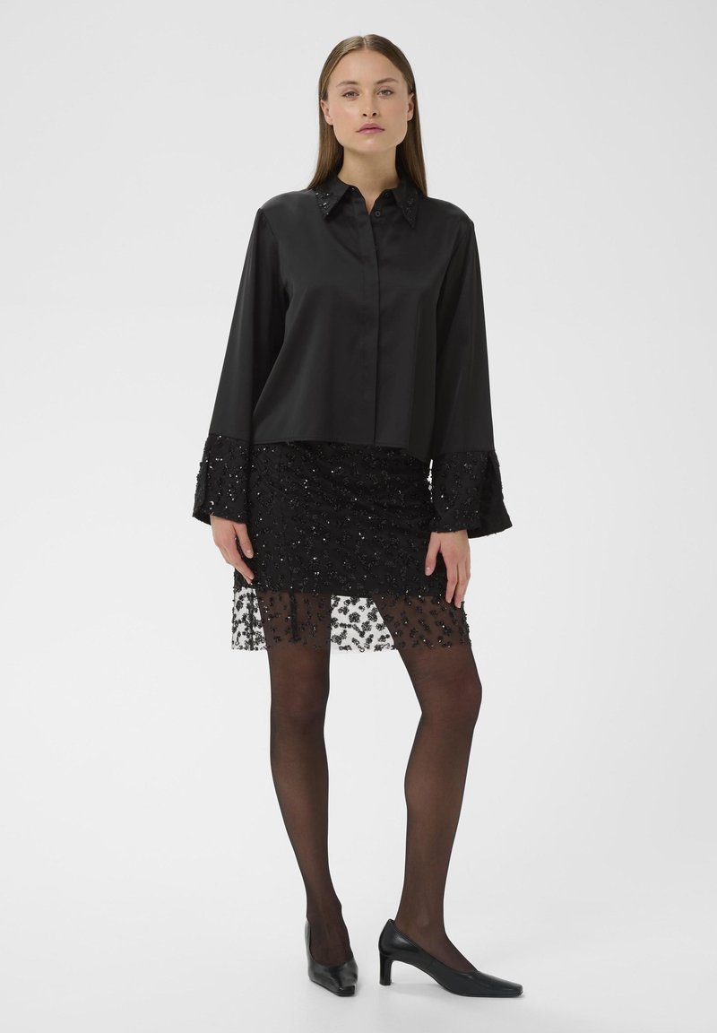 Crna bluzica s oštrim ovratnikom i dugim rukavima, u kombinaciji s crnom suknjom ukrašenom perlicama koja ima čipkastu nadogradnju i crnim tajicama, upotpunjena visokim potpeticama.