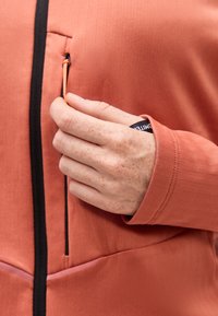 Χέρι με ελαφριές φακίδες τραβάει το πορτοκαλί φερμουάρ σε μπουφάν σκουριάς με μαύρη επένδυση και άνοιγμα τσέπης.