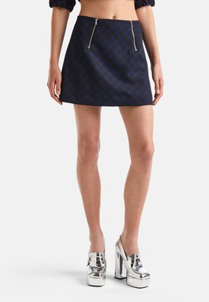 Femme portant une mini-jupe à carreaux bleu marine avec deux fermetures éclair à l'avant et des chaussures à plateforme à talons hauts argentées sur fond blanc.