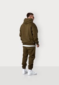 Olive green sweatshirt and matching joggers with ribbed cuffs. White t-shirt underneath. White sneakers with Nike logo. Plain background.