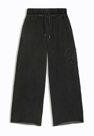 Jonge man met krullend haar, zwarte zonnebril, een donker oversized jas, wijd uitlopende broek en zwarte schoenen, staand tegen een effen achtergrond.