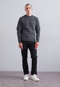 Uomo in piedi contro un muro neutro, con una felpa grigia scura, pantaloni neri e sneaker bianche, espressione neutra e postura rilassata.
