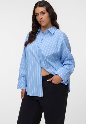 Femme aux longs cheveux foncés portant une chemise ample à rayures bleues et blanches boutonnée et un pantalon noir, posant avec une main dans la poche.
