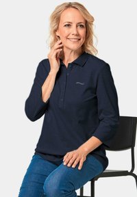 Marineblaues Polohemd mit einer Knopfleiste mit drei Knöpfen, langen Ärmeln und einem kleinen silbernen Logo auf der Brust, kombiniert mit blauen Jeans.