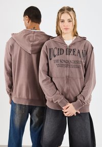 Barna kapucnis pulóver elülső zippel, elől nyomtatott grafikák "LUCID DREAM" és "SONORAN DESERT" felirattal, bordázott mandzsettákkal és aljjal.