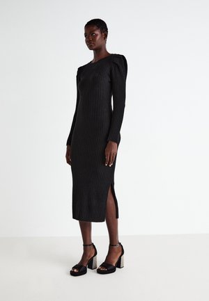 Robe midi noire côtelée à manches longues et fente latérale, présentant une silhouette ajustée. Associée à des sandales à talons noires avec des accents texturés.
