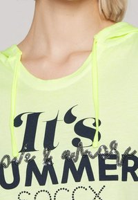 Sweat à capuche vert clair avec des textes en sequins noirs et argentés. Dispose de cordons de serrage et d'une coupe décontractée. Le texte indique : « C'est l'été. »