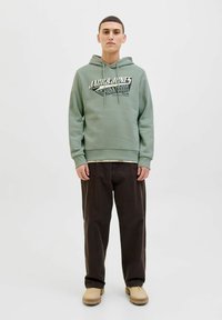 Sweatshirt à capuche vert clair avec un logo graphique, associé à un pantalon ample marron foncé et des chaussures beige. Design simple, look décontracté.