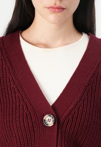 Bordeaux gebreide cardigan met een diepe V-hals en een grote ronde knoop. Gelaagd over een textuur crème kleurig top.