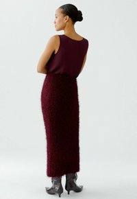Femme aux cheveux bruns en chignon, portant un haut sans manches bordeaux, une jupe midi fuzzy bordeaux et des bottes à talons noirs brillantes, se tenant de côté.