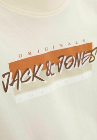 Světlé krémové tričko s barevným potiskem, na kterém je nápis "JACK & JONES" hnědou a bílou barvou, a text "ORIGINALS" nad ním. Vyrobeno z jemné bavlny.