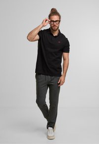 Polo noir à manches courtes avec logo rouge, associé à un pantalon de survêtement gris et des baskets blanches. Le modèle se tient avec des lunettes dans une pose détendue.