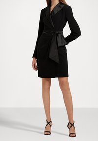 Černé zavinovací šaty se saténovým límečkem a sladěným páskem, dlouhými rukávy a délkově ke kolenům. Nosené s černými sandály na vysokém podpatku.