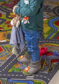 Tout-petit debout sur un tapis de jeu coloré à thème urbain, portant des chaussons renard, un jean bleu et tenant une peluche attachée à une couverture bleue.