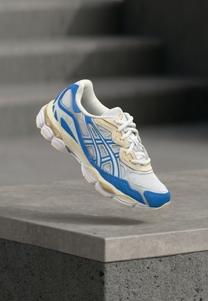 Baltos ir mėlynos sportinės sneakersai su smėlio spalvos akcentais, padėti virš betono krašto, fone matomi neaiškūs pilki laiptai.