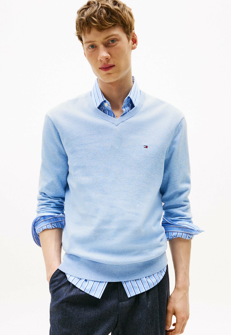 Młody mężczyzna z potarganymi włosami, ubrany w jasnoniebieski sweter w serek na paskowanym koszuli z guzikami, z rękami w kieszeniach, na jednolitym tle.