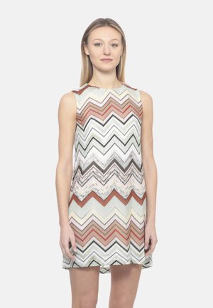 Donna che indossa un vestito senza maniche, lungo fino al ginocchio, con un motivo a zigzag nei toni di marrone, bianco, nero e beige, in piedi davanti a uno sfondo bianco semplice.