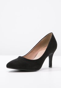 Scarpe con tacco alto da donna in suede nero con punta appuntita e tacco sottile, esposte su una superficie bianca contro uno sfondo grigio chiaro.