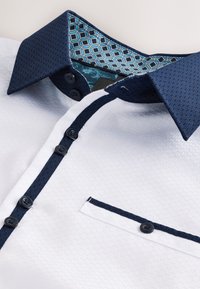 Camicia bianca con tessuto strutturato, colletto blu navy e rifinitura della tasca con motivo geometrico in teal. Sei bottoni scuri sul davanti.