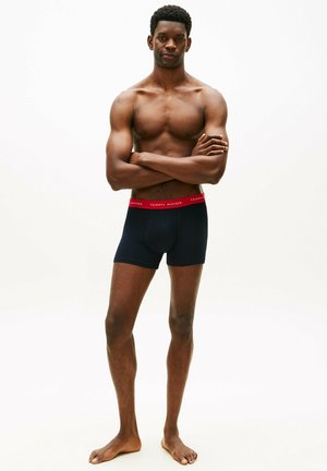 Homme torse nu debout pieds nus, bras croisés, portant des boxer noirs avec une ceinture rouge Tommy Hilfiger, sur un fond blanc uni.