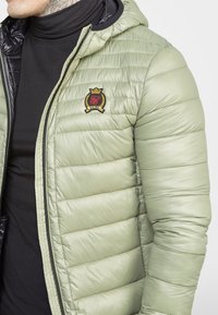 Close-up of a person wearing a light green quilted jacket with a black emblem on the chest over a black turtleneck.