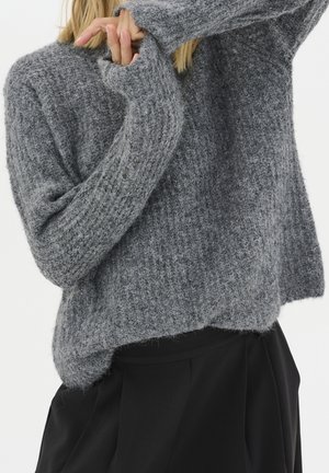 Personne portant un pull en maille gris texturé ample à manches longues et un pantalon noir, les bras partiellement levés couvrant une partie du visage.
