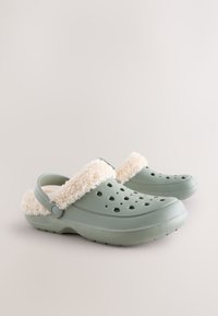 Gröna träskor gjorda av flexibelt material, med en gräddfärgad, fluffig foder, ventilationshål och justerbart hälband.