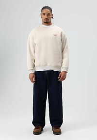 Beige sweatshirt met rode borduursels op de borst, gecombineerd met loszittende donkerblauwe broek en bruine schoenen met rode accenten.