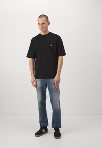 Camiseta de algodón negra con cuello redondo y mangas cortas, que presenta un pequeño logo blanco. Combinada con unos vaqueros azul desgastados y zapatillas negras.
