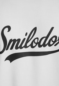 Biała bawełniana tkanina z dużym, wyraźnym czarnym nadrukiem słowa "Smilodos" w stylizowanym kaligraficznym kroju pisma z falistym akcentem.