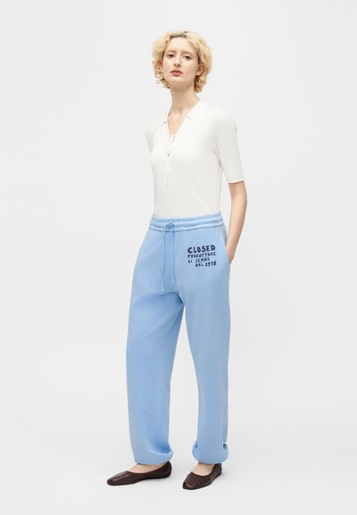 Νεαρός με κοντά ξανθά μαλλιά που φοράει λευκό κοντομάνικο τοπ, ανοιχτό μπλε φόρμες με κείμενο στον μηρό και σκούρα μπαλέτα.