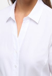 Witte button-up blouse met een gestructureerde stof, gespreide kraag en een v-halsopening met drie zichtbare knopen aan de voorkant.