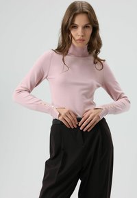 Lichtroze coltrui met lange mouwen, voorzien van decoratieve gouden knopen op de manchetten, gecombineerd met zwarte, hoge taillebroeken.