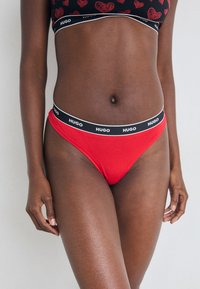 Czerwone majtki bikini z czarnym elastycznym pasem w talii z białym logo "HUGO". Gładki materiał bawełniany, niski krój i minimalne zakrycie.