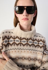 Pull en mailles beige avec un col haut présentant des motifs géométriques marron et blanc. Lunettes de soleil noires avec une monture grise.