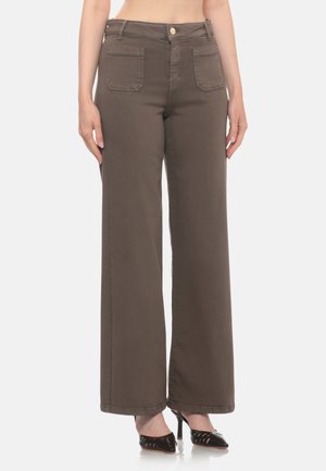 Donna con pantaloni marroni a vita alta e gamba larga con tasche applicate davanti e scarpe nere con tacco alto a punta, in piedi su sfondo bianco.