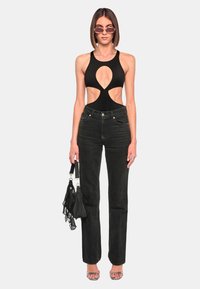 Zwarte badpak met uitsneden, gecombineerd met hoge taille zwarte jeans. Model houdt een kleine zwarte handtas vast en draagt een zonnebril.