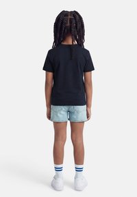 Παιδί με πλεγμένα μαλλιά στέκεται με την πλάτη φανερή, φοράει μαύρο t-shirt, ανοιχτόχρωμο τζιν σορτς, λευκές κάλτσες με μπλε ρίγες και λευκά αθλητικά παπούτσια.