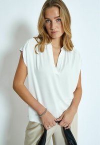 Blusa bianca a maniche corte con scollatura a V, in tessuto morbido, vestibilità rilassata e delicate arricciature sulle spalle, abbinata a pantaloni beige.