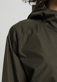 Olivgrön vindjacka med hög krage, slät textur och synliga sömnadsdetaljer på axeln och ärmen.