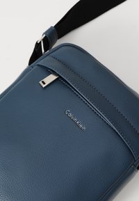 Borsa in pelle blu di Calvin Klein con una texture liscia, forma rettangolare, hardware in argento, tasca con zip e dettaglio del logo sulla parte anteriore.