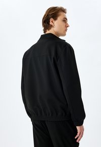 Veste noire à la coupe décontractée, texture lisse, ourlet et poignets élastiqués, et col classique. Design minimaliste sans motifs visibles.