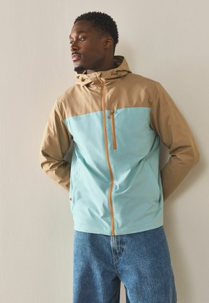 Mann trägt eine beige-hellblaue Reißverschluss-Kapuzenjacke und blaue Jeans, steht mit den Händen hinter dem Rücken vor einer beigen Wand.