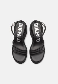 Black strappy sandals with a smooth texture, featuring a wide strap labeled “BUFFALO” and an adjustable ankle strap. Flat sole design.
