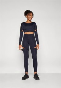 Tengerészkék sportösszeállítás, amely egy kivágásokkal díszített, rövidített hosszú ujjú felsőből és oldalán slisszel ellátott magas derekú leggingsből áll. Sima anyag és testhezálló kialakítás.