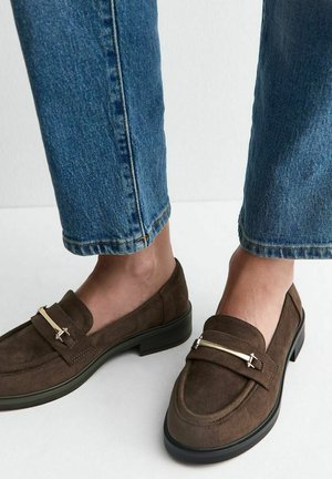 Bruine suède loafers met goud metalen strook detail gedragen met blauwe denim jeans tegen een lichte achtergrond.