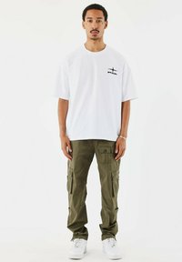 Witte t-shirt met korte mouwen en een zwart logo op de linkerborst, gecombineerd met olijfgroene cargobroek met meerdere zakken en een relaxte fit. Witte sneakers.